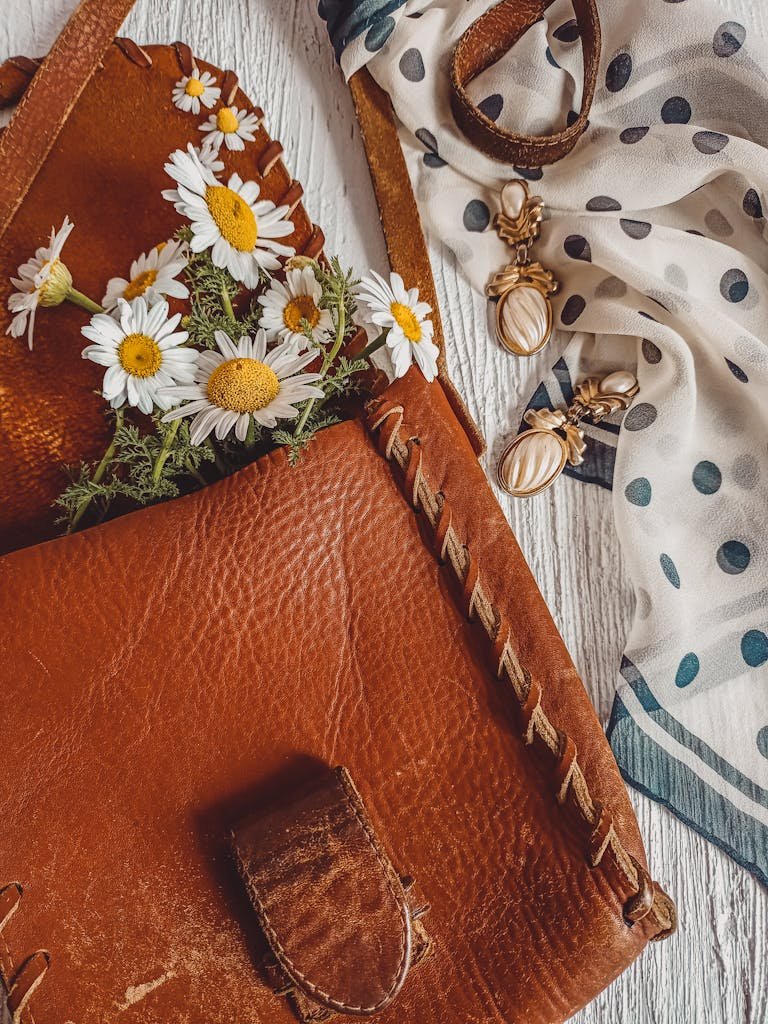 Charming arrangement of a leather bag with daisies and jewelry on a wooden surface.